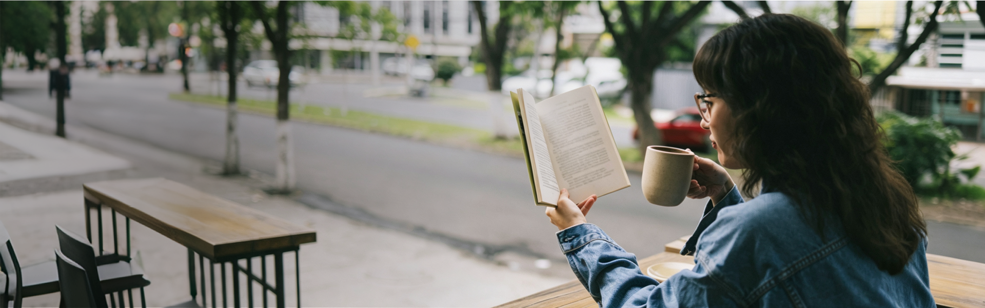 A woman wearing glasses reading a book and drinking coffee at an outdoor cafe